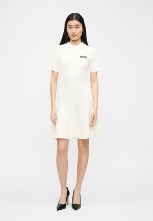 O femeie stă cu fața către înainte, purtând o rochie polo albă cu mâneci scurte și logo-ul "Moschino" și pantofi cu toc înalt negri pe un fundal simplu.