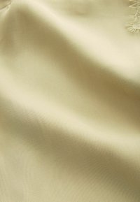 Tissu beige clair et lisse avec un pli subtil et une texture douce, présentant une petite section de franges décoratives dans le coin supérieur droit.