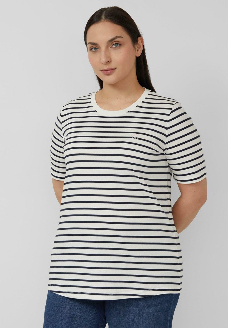 Femme aux longs cheveux foncés portant un t-shirt à manches courtes à rayures horizontales blanches et noires et un jean bleu, debout devant un fond uni.