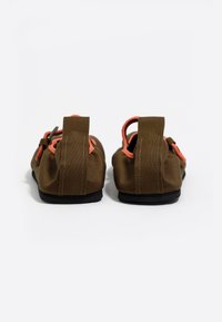 Zapatos de tela marrones con acentos naranjas, con un diseño de perfil bajo y elasticidad, y suelas de goma negra. La vista posterior muestra las lengüetas de agarre.