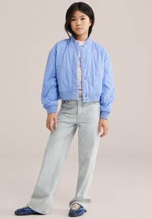 Dekle stoji, oblečena v svetlo modro prešito jakno, cvetlično srajco, svetlo modre kavbojke in modre superge na enobarvnem ozadju.