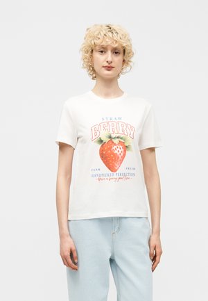 Woman with short curly blonde hair wearing white t-shirt with large strawberry graphic and text, paired with light blue jeans, standing straight.