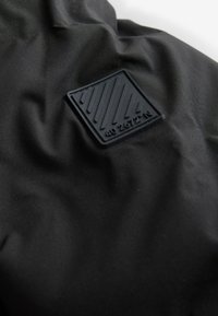 Schwarzer Stoff mit einem strukturierten Gummipatch mit schrägen Linien und den Koordinaten "40 2672° N." Glatte, matte Oberfläche mit minimalem Glanz.