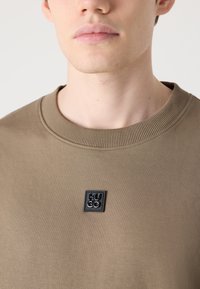 Kaki sweatshirt s koničastim izrezom, opremljenim z rebrasto obrobo, ki ima črn pravokotni značko s reliefnimi črkami na levi strani prsnega koša. Gladka tkanina.