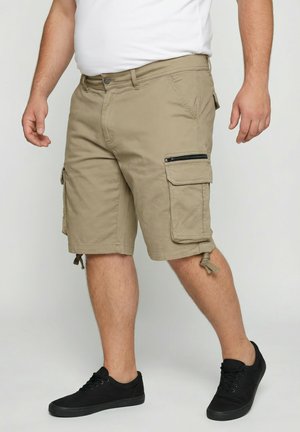 Mężczyzna w beżowych szortach typu cargo z kieszeniami na zamek i patką, czarnych sportowych butach i białej koszulce, stojący na jednolitym tle.