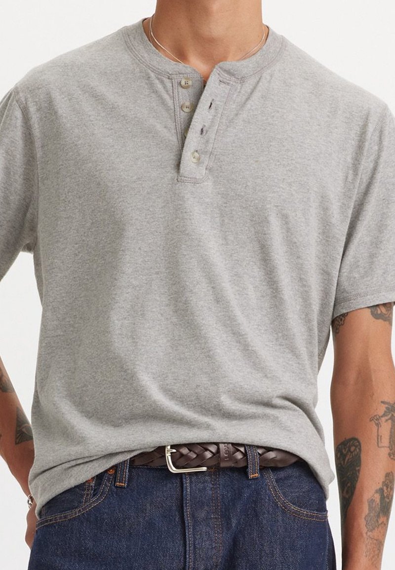 Homme portant un t-shirt henley à manches courtes gris clair, un jean bleu foncé et une ceinture tressée marron avec des tatouages visibles sur les avant-bras.
