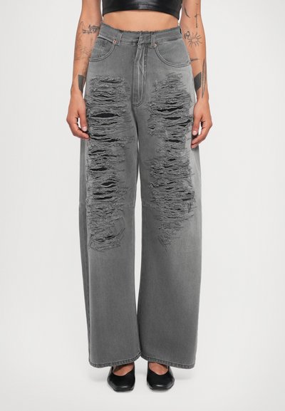 Jean large gris avec des déchirures prononcées sur les deux jambes, taille haute et texture en denim lisse. Des chaussures noires complètent le look.