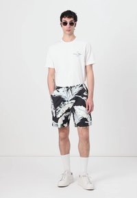 T-shirt bianca, pantaloni corti neri con motivo di foglie di palma bianche e blu, abbinati a sneakers bianche e occhiali da sole. Il modello è in piedi contro uno sfondo semplice.