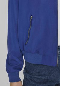 Chaqueta bombera azul con puños y dobladillo acanalados, con un bolsillo con cremallera en el lado izquierdo, hecha de un tejido suave y ligero.