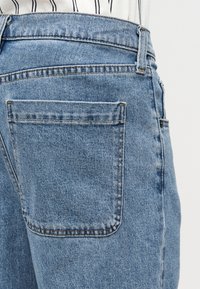 Κοντινό πλάνο της πίσω τσέπης φωτεινού γαλάζιου τζιν με εμφανή ραφές και θηλιές για ζώνη, φορεμένα με άσπρο πουκάμισο με μαύρες ρίγες.