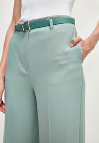 Světle zelené kalhoty s širokými nohavicemi vyrobené z texturované látky, s pevným tyrkysovým páskem se zlatou sponou a diskrétními bočními kapsami.