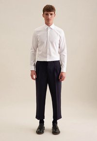 Camicia bianca a maniche lunghe con colletto a punta, abbinata a pantaloni plissettati blu scuro e scarpe nere, in piedi contro uno sfondo uniforme.