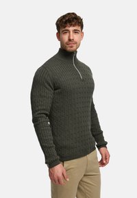 Ciemnozielony, dziergany sweter z wysokim kołnierzem i suwakiem do ćwiartki. Posiada fakturę w formie wzoru warkoczowego i prążkowane mankiety, zestawiony z jasnokawowymi spodniami khaki.