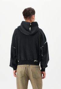 Black cropped hoodie with a distressed design, featuring white paint splatters, a large hood, and loose sleeves. Paired with light tan jeans.
