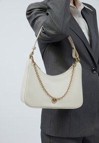 Krämfärgad handväska med en slät läderstruktur, kurvad form, detalj av guldkedja och en logobrand. Bärs med en grå kostym.