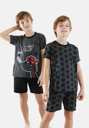 Due ragazzi indossano completi di pigiama coordinati. Il completo a sinistra presenta una maglietta grigia con una grafica di un dinosauro e pantaloni corti neri. Il completo a destra ha un motivo a stelle su sfondo nero.