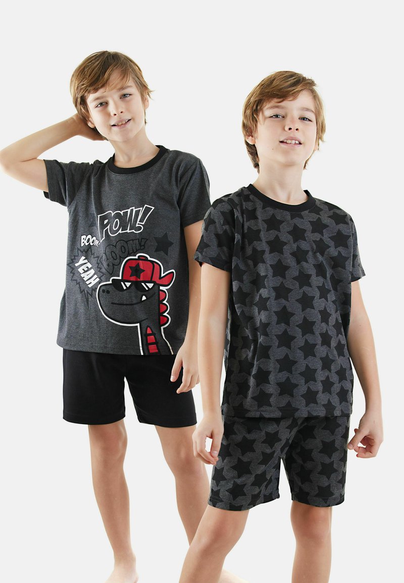 Zwei Jungen tragen passende Schlafanzüge. Das linke Set besteht aus einem grauen T-Shirt mit einem Dinosaurier-Motiv und schwarzen Shorts. Das rechte Set hat ein Sternenmuster auf schwarzem Hintergrund.