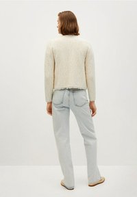 Person mit kurzen braunen Haaren, die einen cremefarbenen strukturierten Pullover und hellblaue, hochgeschnittene Jeans trägt, steht mit dem Rücken zur Kamera.