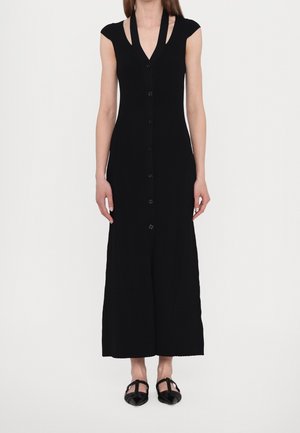 Femme portant une robe maxi noire sans manches boutonnée, col en V, et des chaussures plates noires avec des brides.