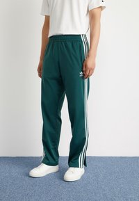 Zelene športne hlače z belimi stranskimi črtami, elastičnim pasom in logotipom blizu bokov. Ujemajo se z belimi supergami na modri preprogi.