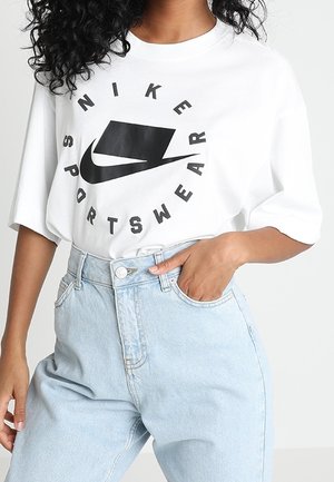 Person iført hvid Nike Sportswear T-shirt, stoppet ned i høj-taljede lyseblå jeans, med den ene hånd i lommen og krøllet hår synligt.