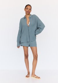 Leichtes, strukturiertes, blau-grünes Hemd mit Knöpfen, kombiniert mit passenden Shorts, in entspannter Passform und mit subtilen Details. Getragen mit Sandalen.