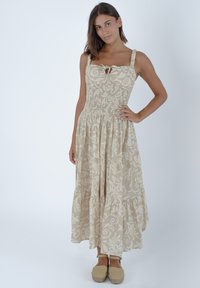 Jasno beżowa sukienka z kwiatowym wzorem, kwadratowym dekoltem i wiązaniem. Posiada marszczoną talię i warstwową spódnicę. Noszona z butami espadrilles.