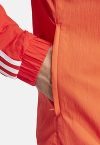 Chaqueta roja con un tejido texturizado, puños elásticos con franjas blancas y bolsillos laterales. El diseño es simple y funcional.