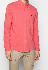 Camicia a maniche lunghe con bottoni di colore corallo, realizzata in tessuto morbido, caratterizzata da un colletto classico e un piccolo logo ricamato sul petto.