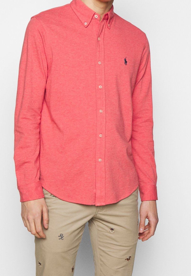 Camicia a maniche lunghe con bottoni di colore corallo, realizzata in tessuto morbido, caratterizzata da un colletto classico e un piccolo logo ricamato sul petto.