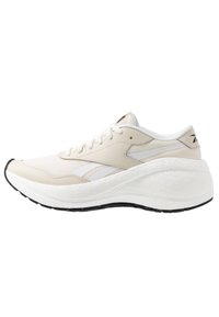 Beige sportskor med vita detaljer, övre i mesh, tjock texturerad mellansula och ett mönstrat svart yttersula. Snörning och minimal logotypdetalj.