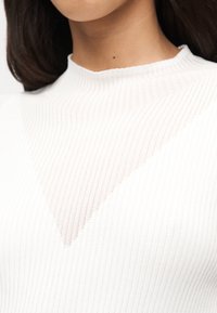 Κοντινό πλάνο ενός ατόμου που φοράει λευκό πλεκτό πουλόβερ με ραβδώσεις, με διαφανές V-σχήματος σχέδιο στο στήθος και ψηλό λαιμό.