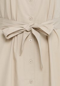 Beige stoffen jurk met knopen aan de voorkant en een ceintuur in de taille die als strik in het midden van het kledingstuk is gebonden.