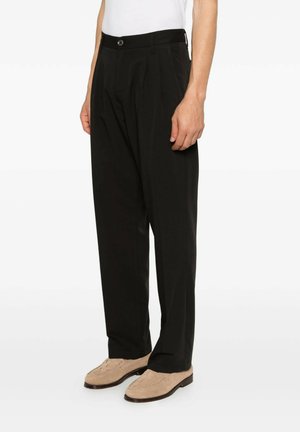 Uomo con pantaloni neri plissettati e mocassini beige in suede, in piedi su sfondo bianco.