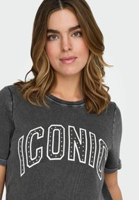 Hall hall t-särk, millel on keritud varrukad ja peal sõna "ICONIC" paksus valges kirjutuses, mis on kaunistatud metallist naeltega, valmistatud pehmest tekstiilist.