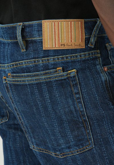 Blugi de denim în albastru închis cu un finisaj texturat. Prezintă o etichetă din piele maro pe talie, cu dungi verticale colorate.