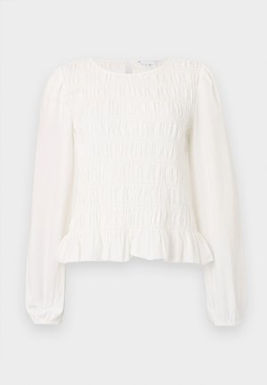 Blouse blanche avec une texture smockée, col rond, manches longues bouffantes et un ourlet froncé à la taille. Matière en coton léger.