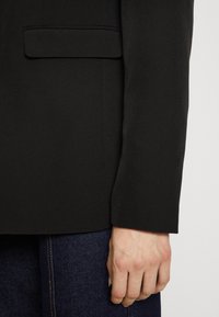 Černý blejzr s hladkým povrchem a jednou náprsní kapsou. Nosit na tmavých džínech, které ukazují čisté linie a přiléhavý střih.