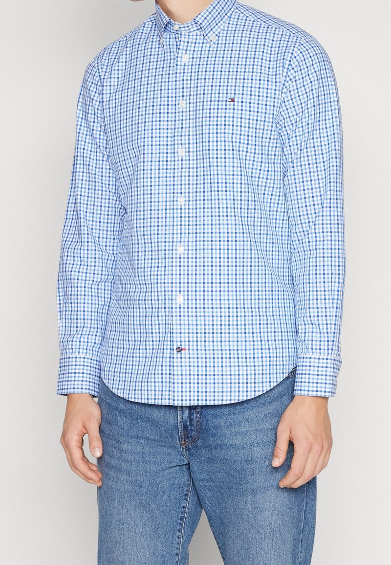 Homme portant une chemise à carreaux bleue et blanche à manches longues boutonnée et un jean bleu, debout devant un fond uni.