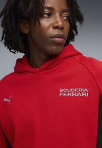 Červená mikina s kapucí a šedým textem "SCUDERIA FERRARI" a černými akcenty. Měkká tkanina s volným střihem, na levé části hrudi se nachází logo Puma.