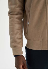 Veste en polaire marron à texture lisse, avec poignets côtelés, poches latérales et boutons-pression. Présente une coupe décontractée et peu de détails.