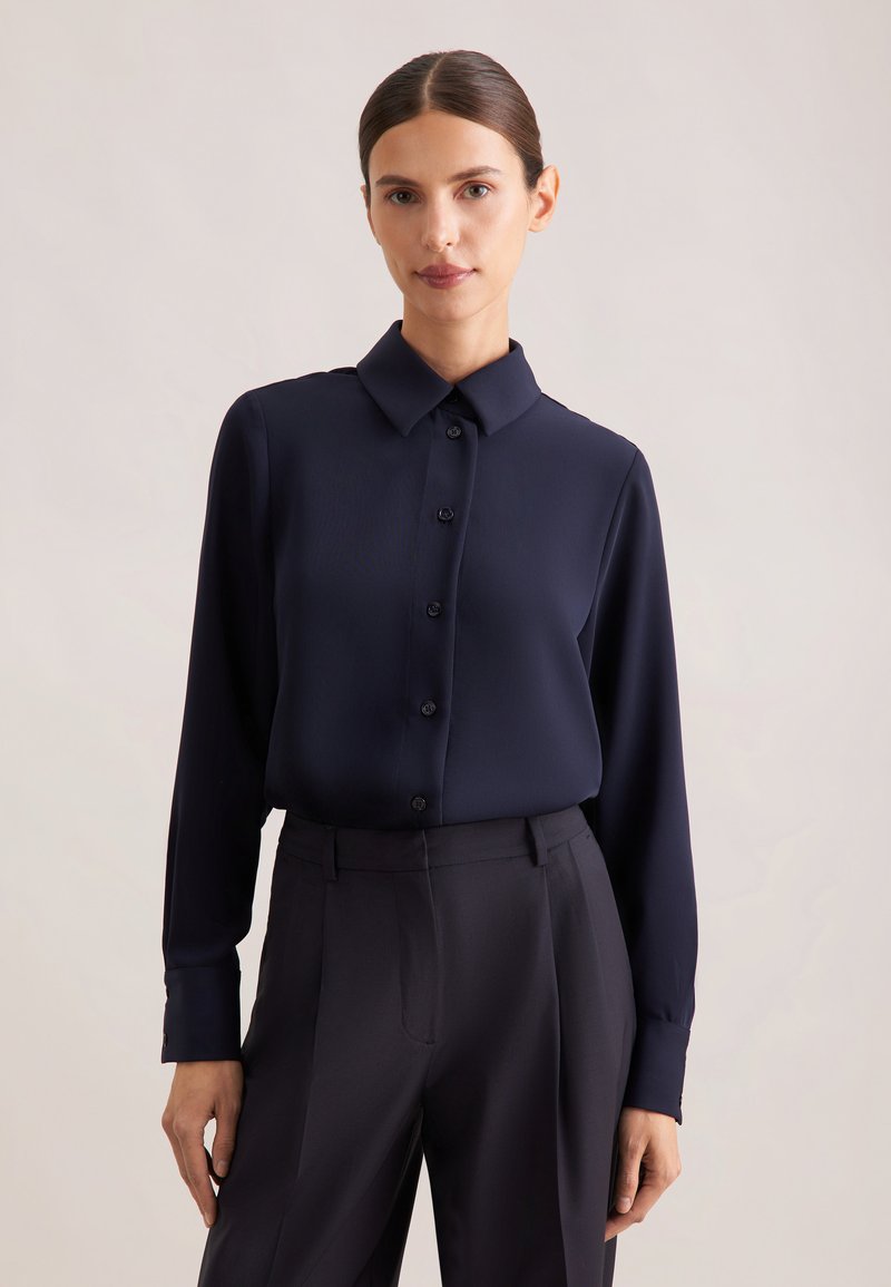 Femme aux cheveux foncés attachés, portant une chemise bleu marine à manches longues boutonnée et un pantalon sombre sur un fond uni.