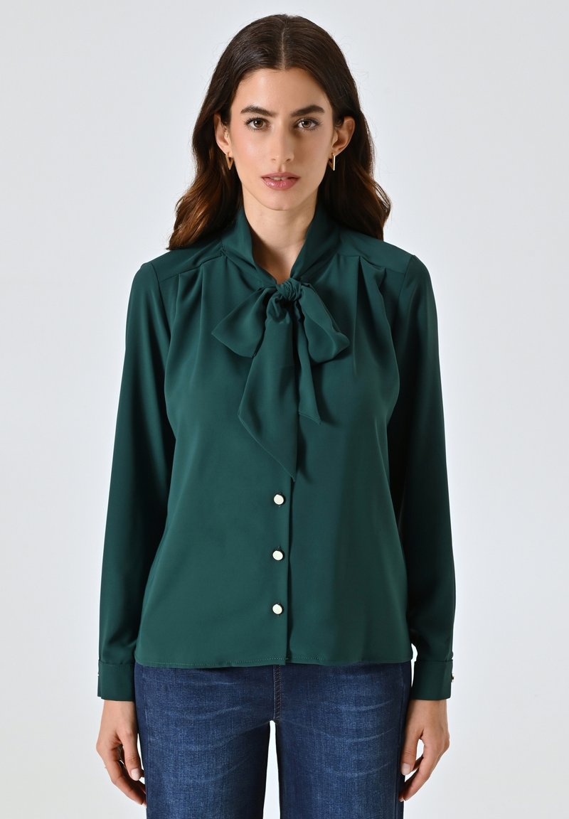 Blusa verde realizzata in tessuto morbido, con scollo a fiocco, maniche lunghe e una fila di cinque bottoni lucidi sul davanti.