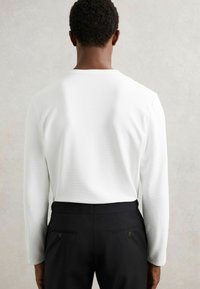 Mann, der sich abgewandt hat, trägt ein schlichtes weißes Langarmhemd, das in schwarze maßgeschneiderte Hosen gesteckt ist, vor einem neutralen Hintergrund.