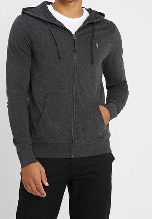 Mężczyzna w ciemnoszarej bluzie z kapturem i zamkiem błyskawicznym, posiadającej przednie kieszenie, na białym t-shircie, stojący z jedną ręką w kieszeni.