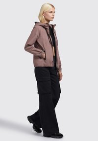 Leichte, mauve Farbe Jacke mit Kapuze und Kordelzügen, ausgestattet mit elastischen Bündchen und Seitentaschen, kombiniert mit schwarzen Cargo-Hosen und schwarzen Schuhen.