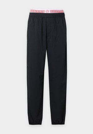 Jeune homme portant un sweat à capuche gris zippé, un t-shirt blanc avec le logo Versace, un pantalon noir et des bottes en cuir noires, debout devant un fond blanc uni.