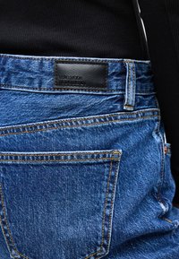 Τσέπη πίσω από μπλε τζιν με πορτοκαλί ραφές και μαύρη δερμάτινη ετικέτα επωνυμίας πάνω από τη μέση, μπροστά από μαύρο ριμπ μπλουζάκι.