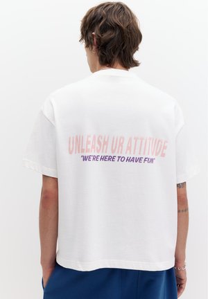 Középhosszú barna hajjal rendelkező személy fehér pólót visel, amelynek hátoldalán az „UNLEASH UR ATTITUDE” és a „WE'RE HERE TO HAVE FUN” felirat szerepel.