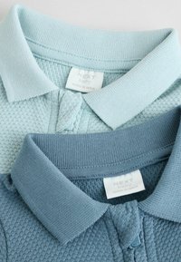 Dos camisetas de polo para bebés en azul claro y verde azulado apagado. La azul clara tiene un tejido texturizado, mientras que la verde azulado cuenta con una tela suave. Etiquetas visibles.